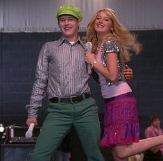 a man standing next to a woman on stage with a microphone in his hand and wearing a green hat