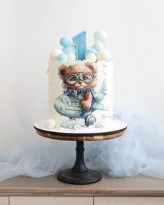 a birthday cake with a teddy bear wearing glasses on it's top and balloons around the edges