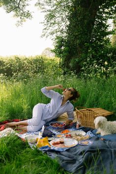 a woman laying on a blanket in the grass next to a picnic basket and dog