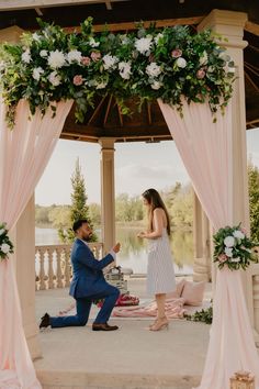 a man kneeling down next to a woman in front of a gazebo with flowers on it