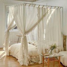 a white canopy bed sitting in a bedroom on top of a hard wood floor next to a window