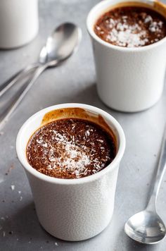 two white cups filled with chocolate pudding on top of a gray table next to spoons