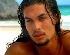 a man with long hair and no shirt on sitting in front of the ocean looking at the camera