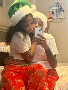 two people sitting on a bed taking a selfie with a cell phone wearing christmas pajamas and santa hats