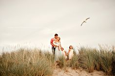 three people are standing in the sand near some tall grass and seagulls flying overhead