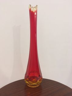 a red glass vase sitting on top of a wooden table next to a white wall