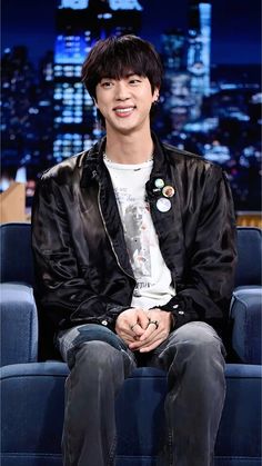 the young man is sitting down in front of a tv screen wearing a black jacket and jeans