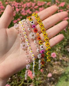 a hand is holding several bracelets with flowers on them in front of some pink and yellow flowers