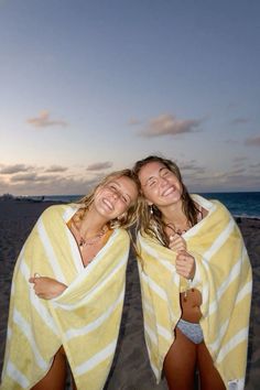 two young women wrapped in towels on the beach at sunset, both smiling and holding their arms around each other