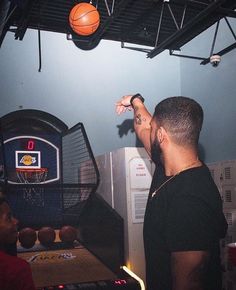 two men are playing basketball in a room with blue walls and black ceiling, while one man is pointing at the hoop