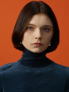 a woman with short brown hair wearing a black turtle neck sweater and earrings, against an orange background