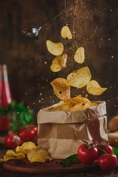 potato chips falling into a bag with tomatoes and other ingredients in the background, on a wooden table