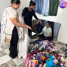 two women standing over a pile of clothes in a room next to a woman sitting on the floor