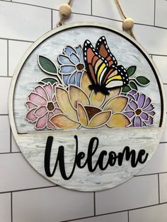 a welcome sign hanging from the side of a brick wall with flowers and a butterfly on it