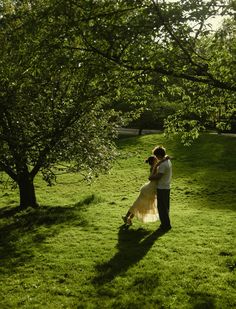 a man and woman are standing in the grass under some trees, hugging each other