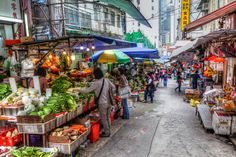 an outdoor market with lots of vegetables and people walking around the storefronts on both sides