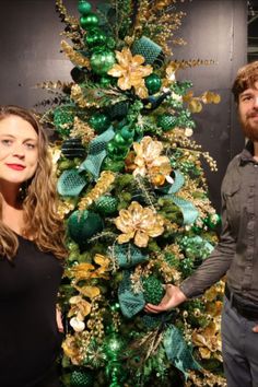 a man and woman standing next to a green christmas tree with gold ornaments on it