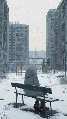 a person sitting on a park bench in the middle of a snowy day with buildings in the background