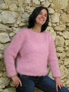 a smiling woman in pink sweater leaning against a stone wall with her hands on her hips