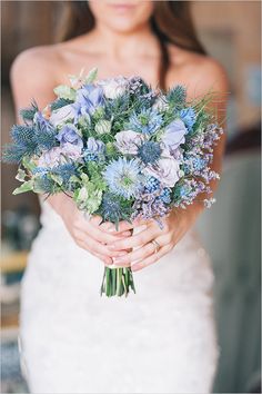 a woman in a wedding dress holding a bouquet of blue and white flowers with greenery