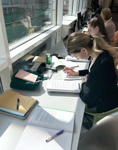 two women sitting at a table with notebooks and books in front of them, looking out the window