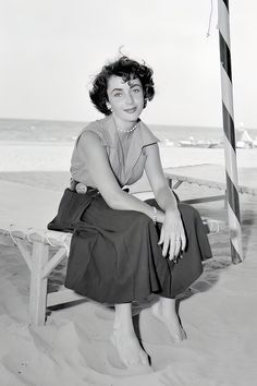 a black and white photo of a woman sitting on a bench in the sand at the beach