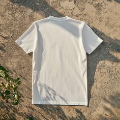 a white t - shirt laying on the ground next to a plant with leaves in it