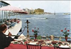 a man sitting at an outdoor table with food and drinks on it overlooking the water