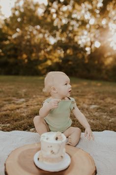 a baby sitting on the ground with a cake in front of it and trees in the background