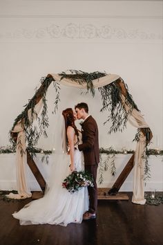 a bride and groom kissing in front of an arch decorated with greenery