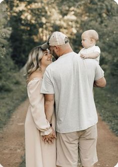 a man and woman holding a baby while standing on a dirt road with trees in the background