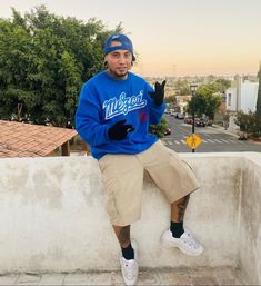 a man sitting on top of a cement wall wearing a blue sweatshirt and khaki shorts