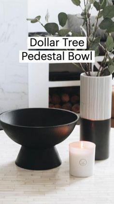 a candle sits on a table next to a black bowl and white vase with greenery in it