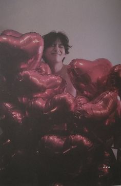 a woman sitting on top of a pile of shiny red heart shaped balloons in front of a white wall