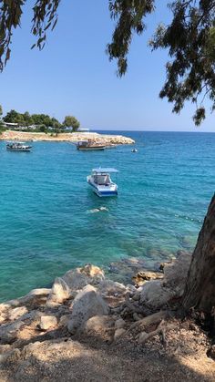 boats are floating in the blue water near some rocks and trees on the shore line