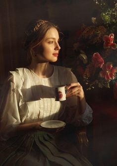 a woman sitting in a chair holding a coffee cup and looking out the window with red flowers behind her