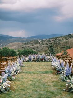 an outdoor ceremony setup with wooden chairs and flowers on the grass in front of mountains