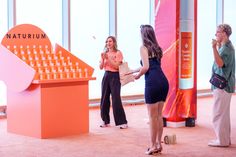 two women standing in front of a display with an orange structure on the floor and people looking at it