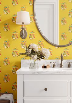 a bathroom with yellow wallpaper and flowers on the counter top, along with a round mirror