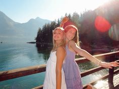 two girls are standing on a bridge near the water and trees, one is holding her arm around the other's shoulder