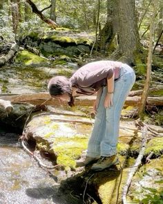 a woman bending over to look at a small stream in the middle of a forest