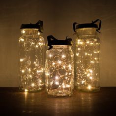 three mason jars filled with fairy lights on top of a wooden table in front of a wall