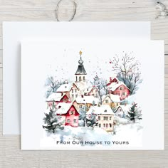 a christmas card with a watercolor painting of houses and trees in the snow, on top of a wooden table