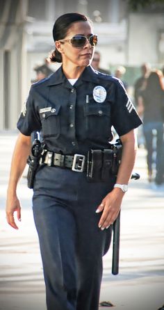 a woman police officer walking down the street with her hand in her pocket and sunglasses on