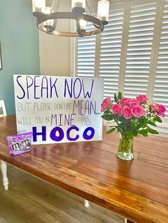 a wooden table topped with a vase filled with pink flowers and a sign that says speak now but please don't be mean