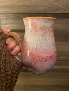 a hand holding a pink and white vase in front of a wooden wall with a brown sweater on it