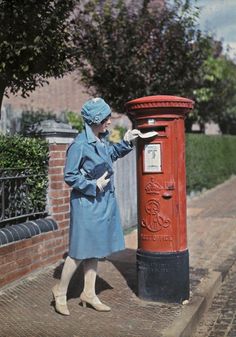 a woman in blue coat standing next to a red mailbox on sidewalk near brick building