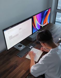 a person sitting at a desk in front of two computer monitors and writing on a notepad