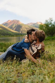 a man and woman kissing in the grass with mountains in the backgrouund