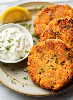 two crab cakes on a plate with a bowl of sour cream and lemon wedges